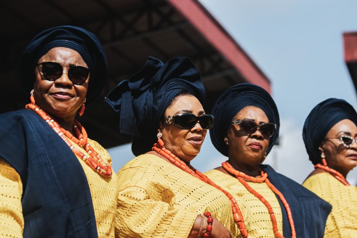 Four women in matching yellow lace tops, navy geles, and coral beads at a Nigerian celebration