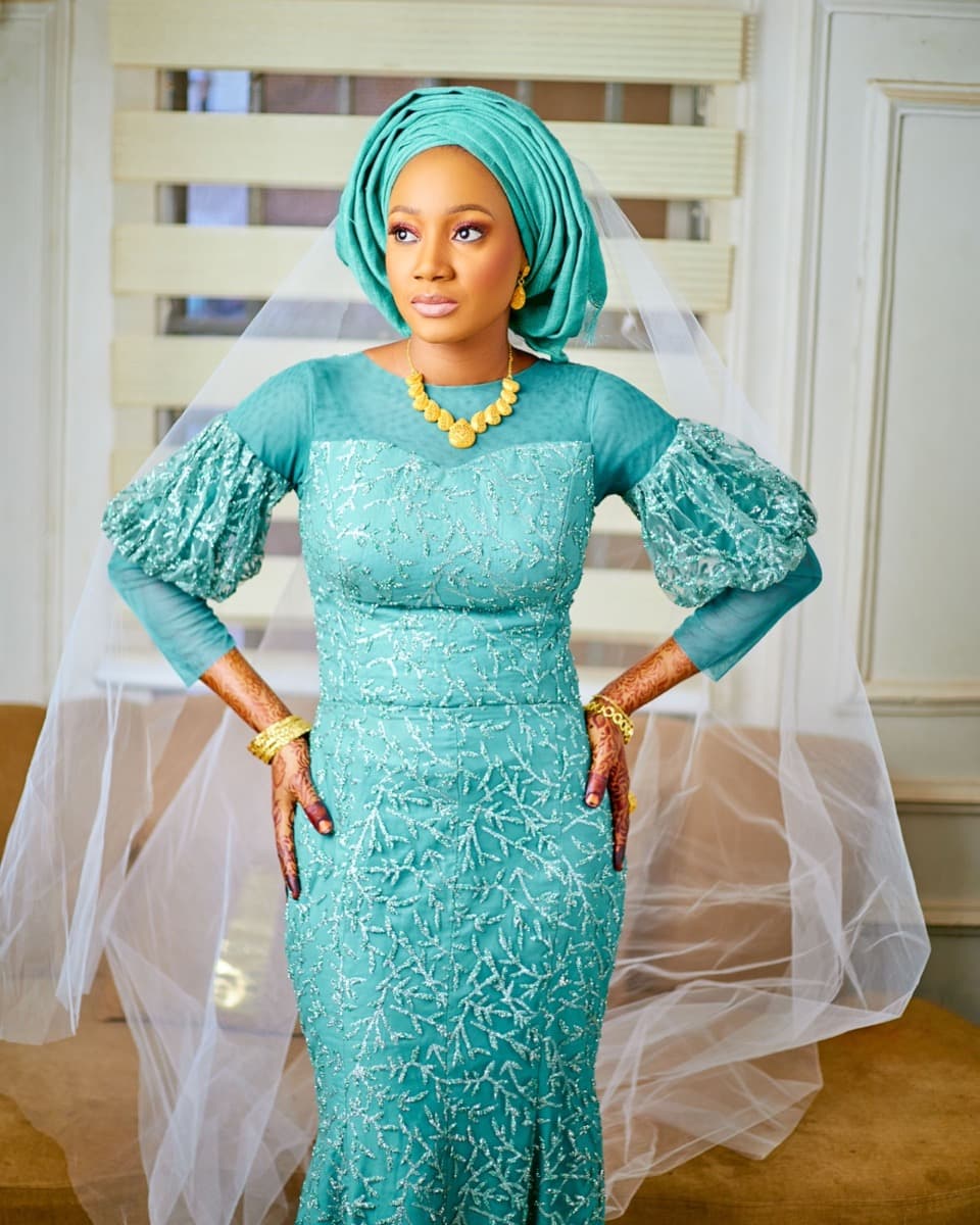 A woman in a fitted teal lace gown with structured puffed sleeves and a matching headwrap