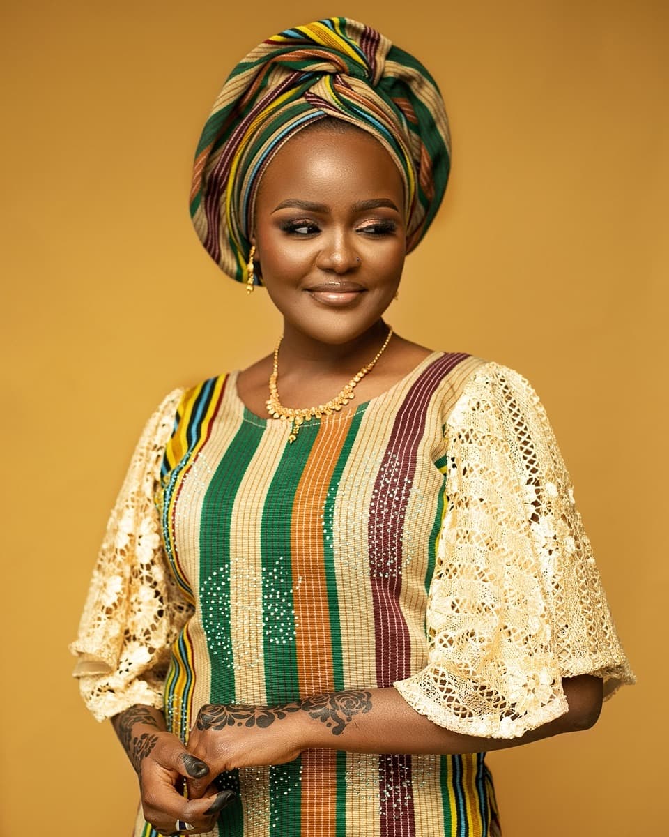 A woman in a green, orange, and maroon striped aso oke top with cream crochet-lace bell sleeves, worn with a matching gele