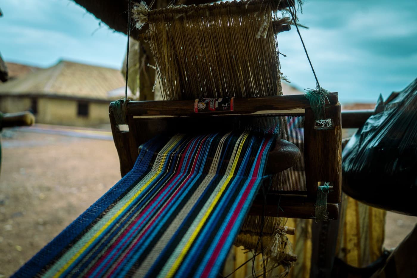 A narrow-strip aso oke loom mid-weave, with blue, yellow, red, and white striped cloth being woven outdoors in a Nigerian village setting