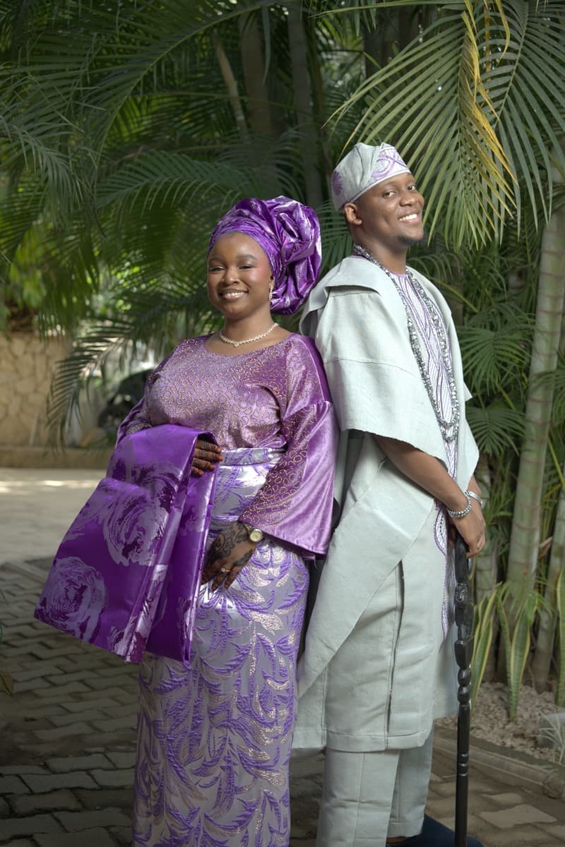 A woman in a shimmery purple glitter buba blouse over a stiffer silver and purple metallic brocade iro wrapper, with a purple gele and draped ipele