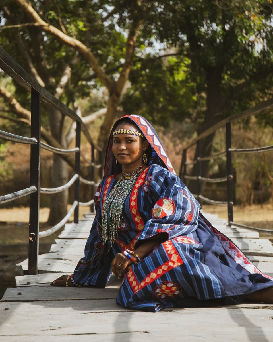 A Fulani woman in an indigo and red patterned robe with a beaded headpiece and layered necklaces, seated on a wooden bridge