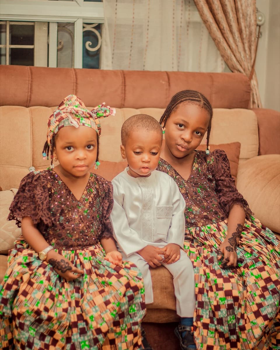 Three siblings on a couch, two girls in matching patterned dresses and a young boy in a white senator-style outfit between them